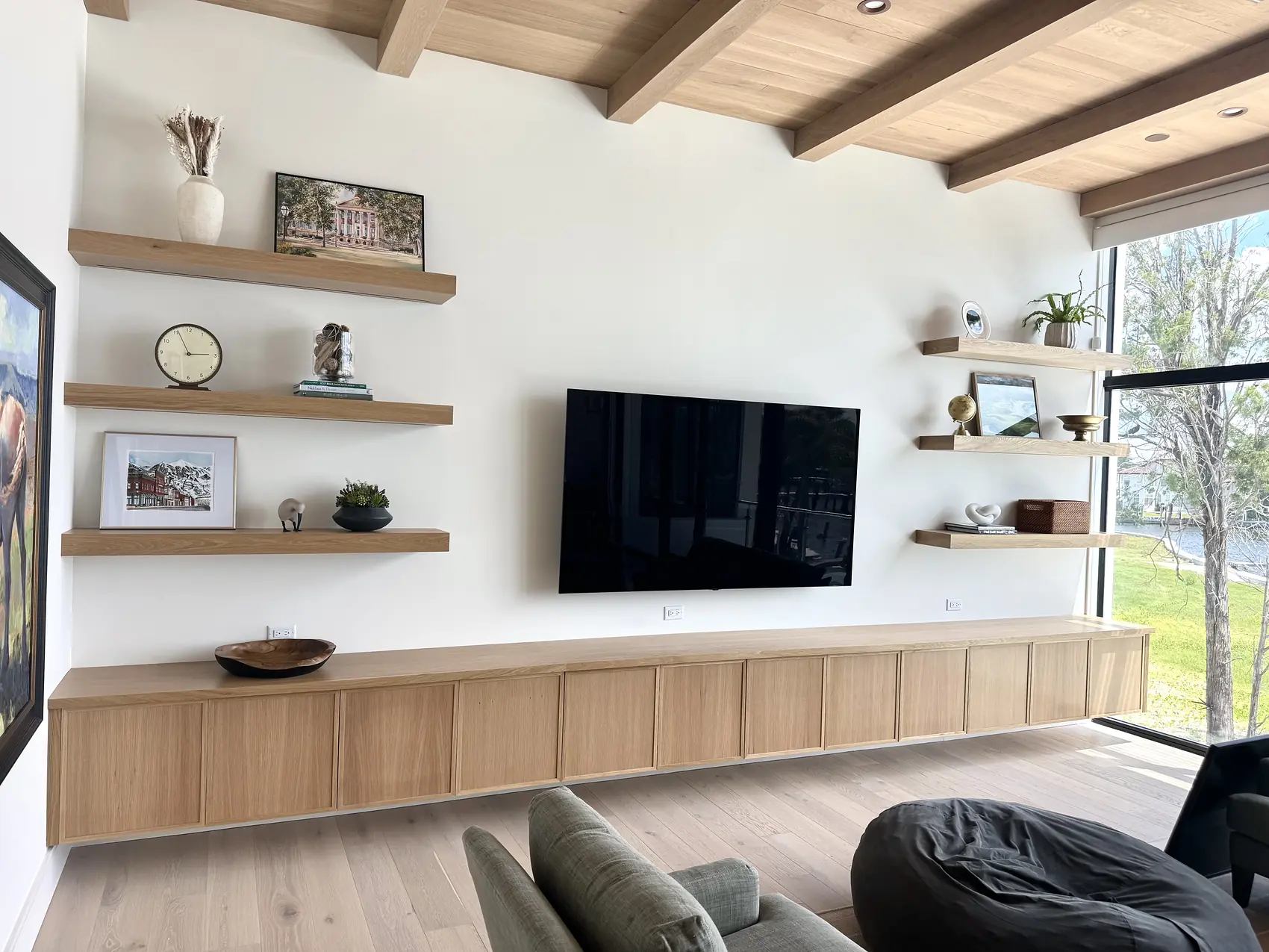 Floating white oak shelves either side of a wall mounted television above a long oak credenza
